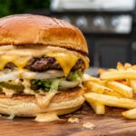 Close-up of a homemade In-N-Out style cheeseburger with melted cheese, creamy burger sauce dripping from toasted buns, crispy fries, and a dipping cup of sauce on a wooden board in a backyard BBQ setting.