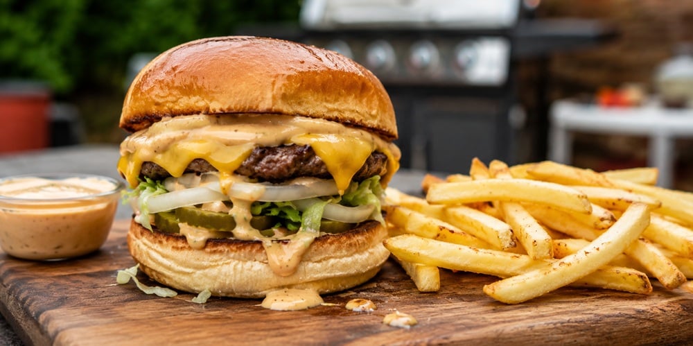 Close-up of a homemade In-N-Out style cheeseburger with melted cheese, creamy burger sauce dripping from toasted buns, crispy fries, and a dipping cup of sauce on a wooden board in a backyard BBQ setting.
